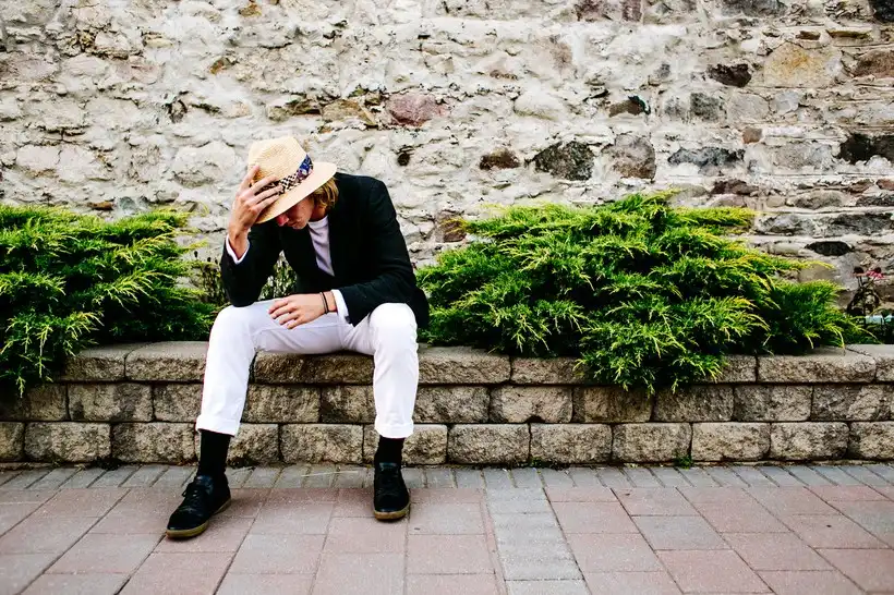 Homme assis en blazer noir, pantalon blanc et chapeau de paille, devant un mur de pierre