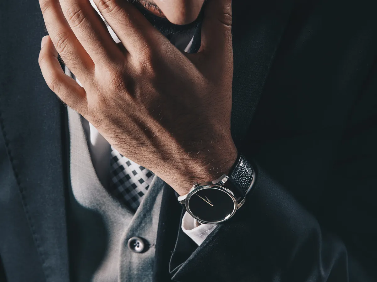Homme en veste de costume sombre sur un gilet gris boutonné, cravate à damier et montre à cadran noir