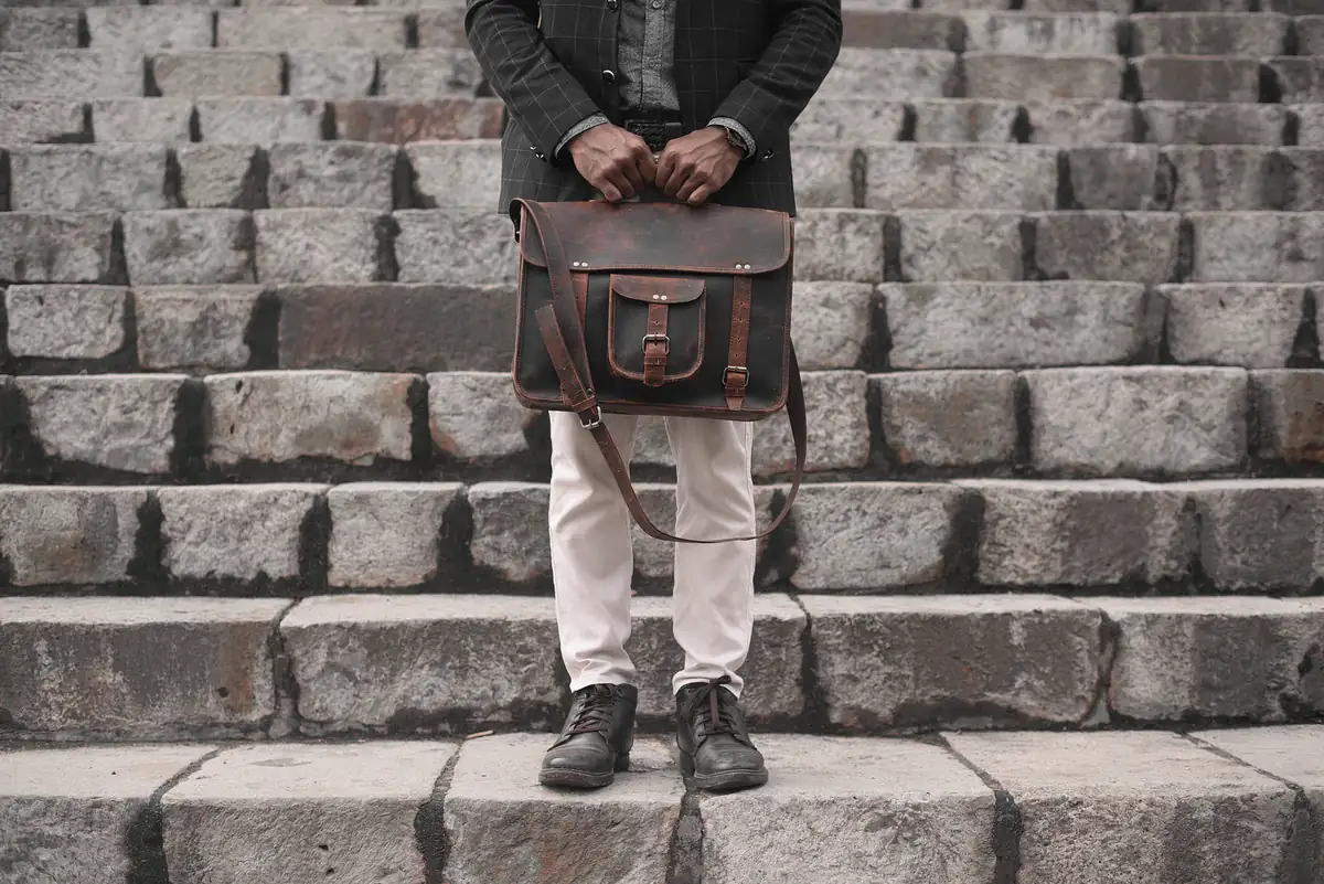 Homme en veste sombre et pantalon clair tenant une besace en cuir brun devant un escalier en pierre