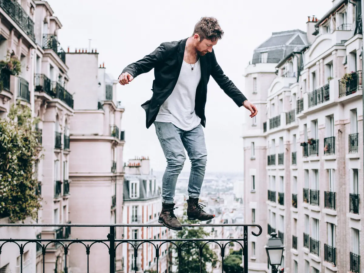 Homme en veste noire, t-shirt blanc, jean délavé et boots marron, sur une rambarde au-dessus des toits de Paris